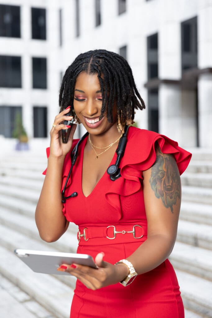nurse wearing red jumpsuit holding ipad in her hand and in the other hand holding a cell phone to her ear while smiling for the camera